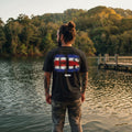 The Beauty Closet black cotton t-shirt with Costa Rica flag graphic on back, worn by a man standing by a lake.