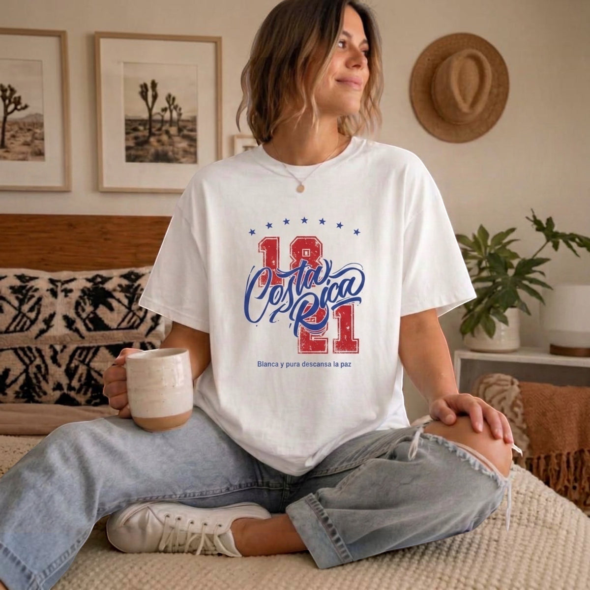 The Beauty Closet cream cotton t-shirt with Costa Rica 1821 graphic print worn by a woman sitting on a bed