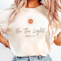 The Beauty Closet cream cotton t-shirt featuring a sun graphic and Be The Light Matthew 5:14 text on a woman