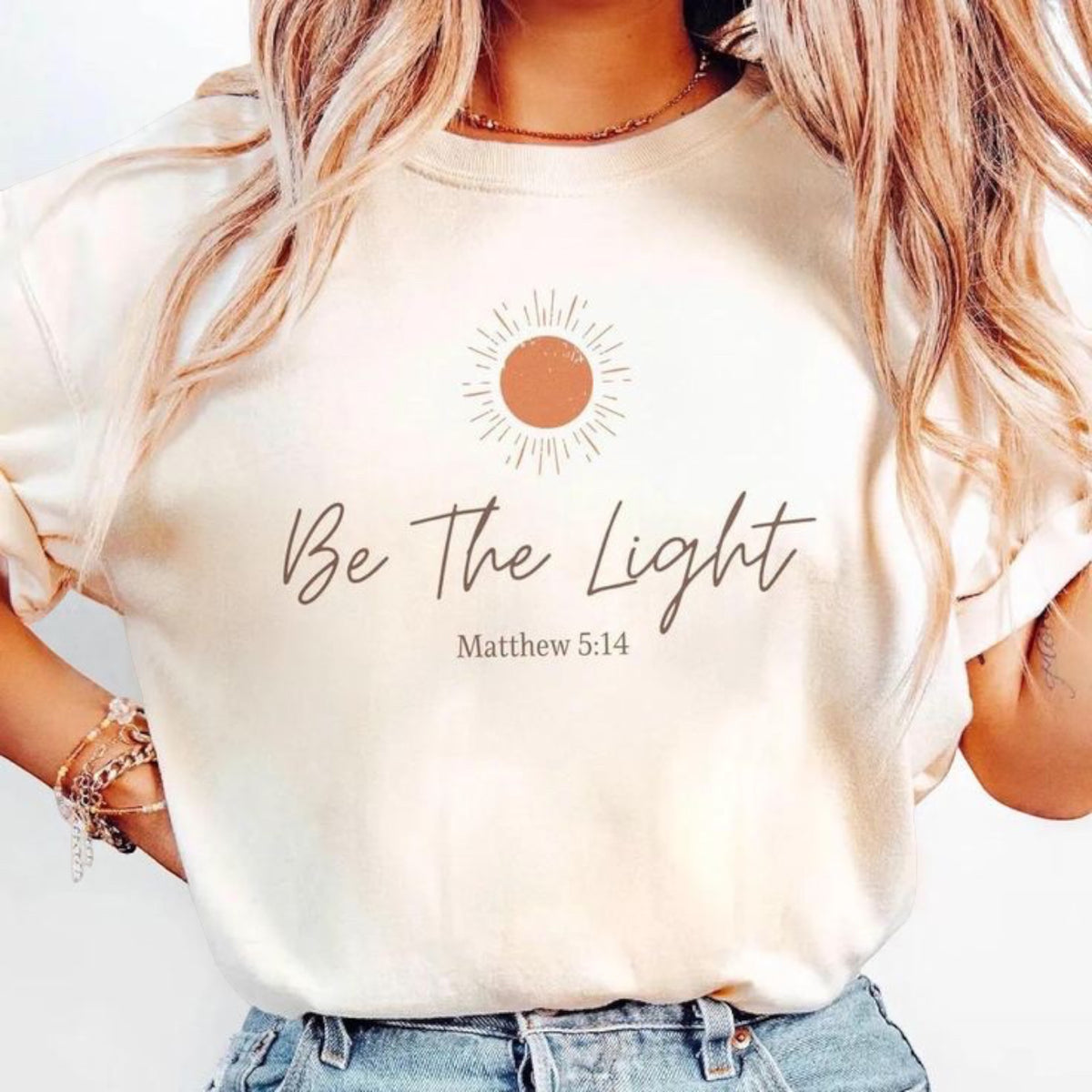 The Beauty Closet cream cotton t-shirt featuring a sun graphic and Be The Light Matthew 5:14 text on a woman