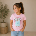 A young girl wearing a light pink The Beauty Closet t-shirt featuring a cute owl graphic with glasses.