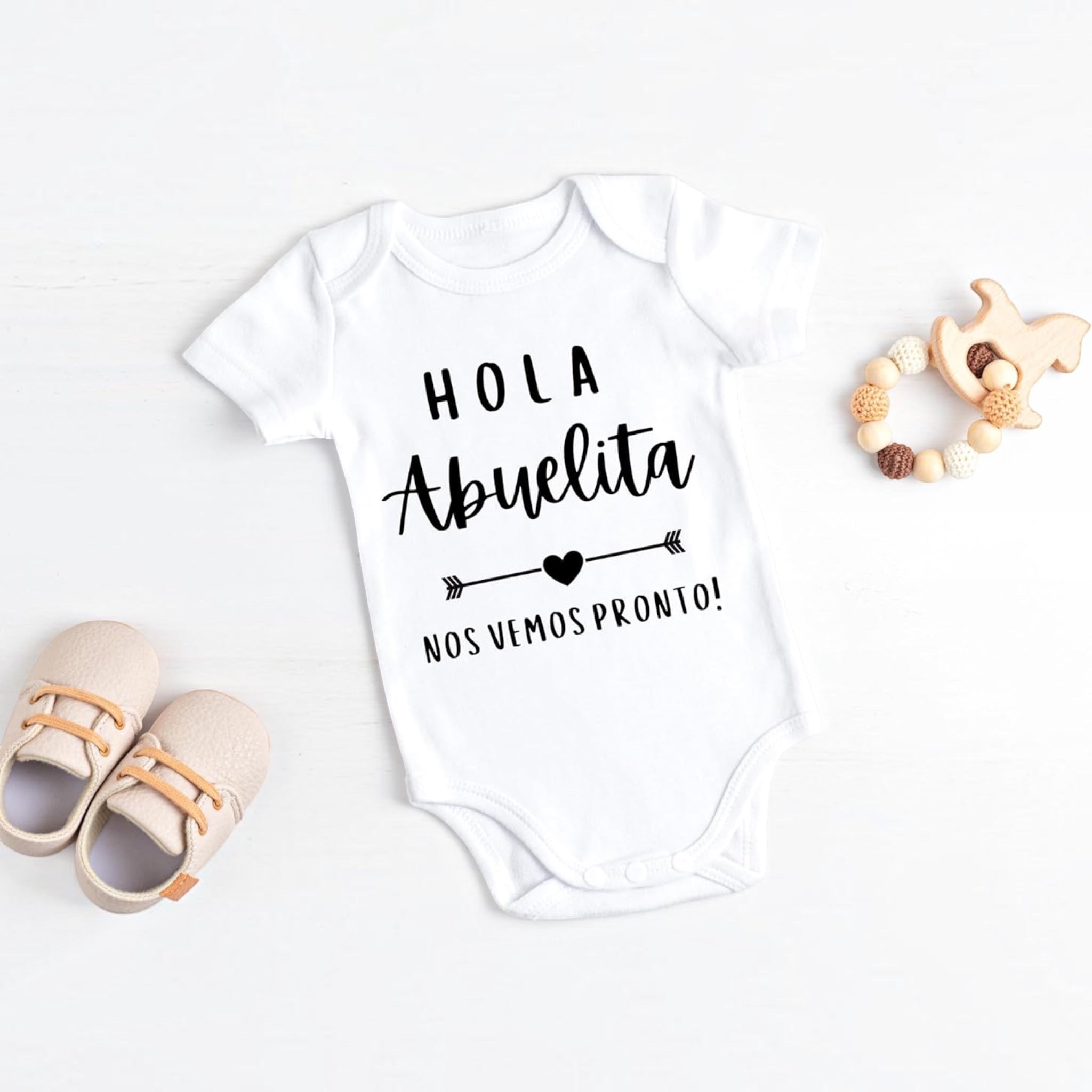 The Beauty Closet white baby bodysuit featuring Hola Abuelita text with a heart and arrow graphic on a wooden surface.